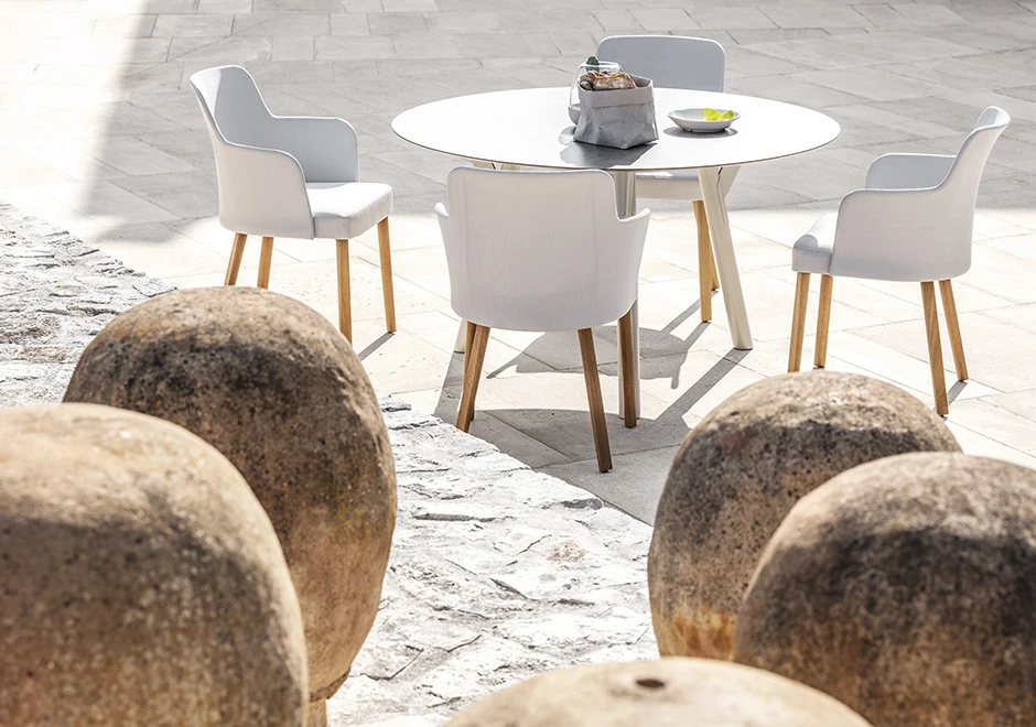 Quatro cadeiras assinadas com estrutura em madeira Iroko natural e assento revestido em tecido bege e encosto com estrutura de tecido. Visto de frente em uma area externa.