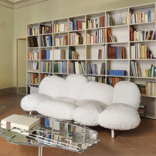 Sofá de design moderno, com estrutura de aço e estofado em pelúcia branco, visto de lado, em uma sala com uma mesa espelhada a frente, um piano ao lado direito e uma estante de livros ao fundo.