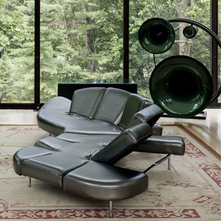 Sofá de design futurista, com estrutura de aço e mecanismo flexível com diferentes inclinações, na cor prata, visto de lado em uma sala moderna com parede de vidro.