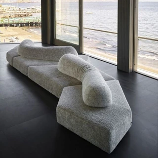 Sofá de design moderno, com pés em ABS, revestido em espuma e com estofado em tecido cinza, visto de lado em uma sala de parede de vidro.