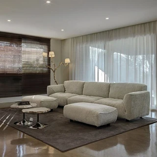 Sofá modular com encosto articulado e estofado em tecido creme, visto de lado, em uma sala aconchegante.