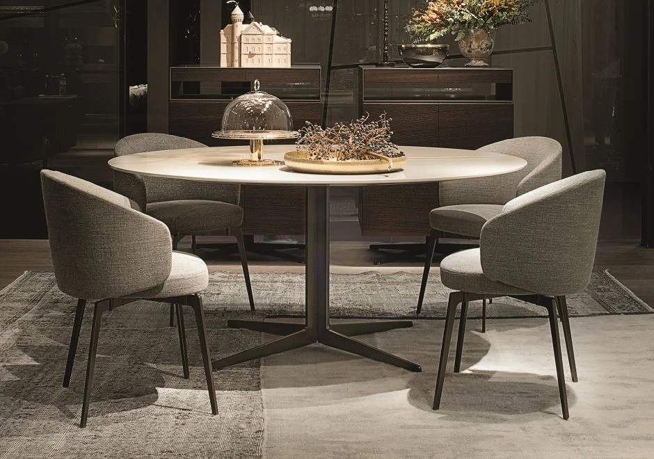quatro cadeiras assinadas estofadas cinzas em volta de uma mesa redonda, com dois objetos de decoração sobre ela. Dois móveis de madeira ao fundo com objetos de decoração sobre eles.