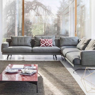 Sofá modular, com estrutura de metal, estofado em tecido cinza e em formato L, visto de frente em uma sala de parede de vidro.