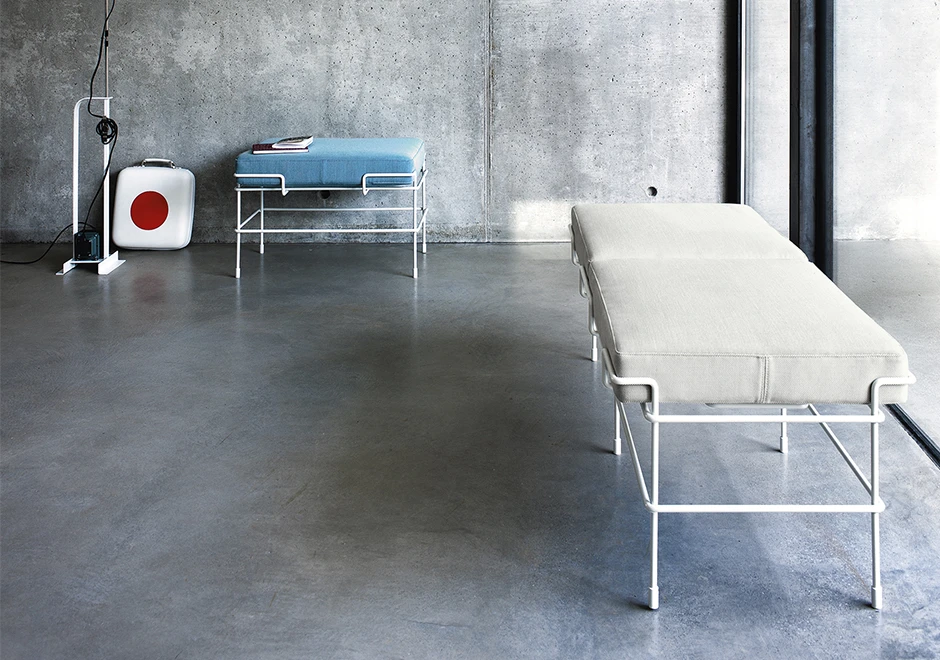 Banco de design com estrutura de aço pintado e almofada de poliuretano expandido com capa de tecido ou couro branco, visto de lado em uma sala vazia.