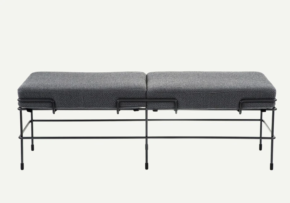Banco de design com estrutura de aço pintado e almofada de poliuretano expandido com capa de tecido ou couro cinza, visto de frente, em um fundo branco.