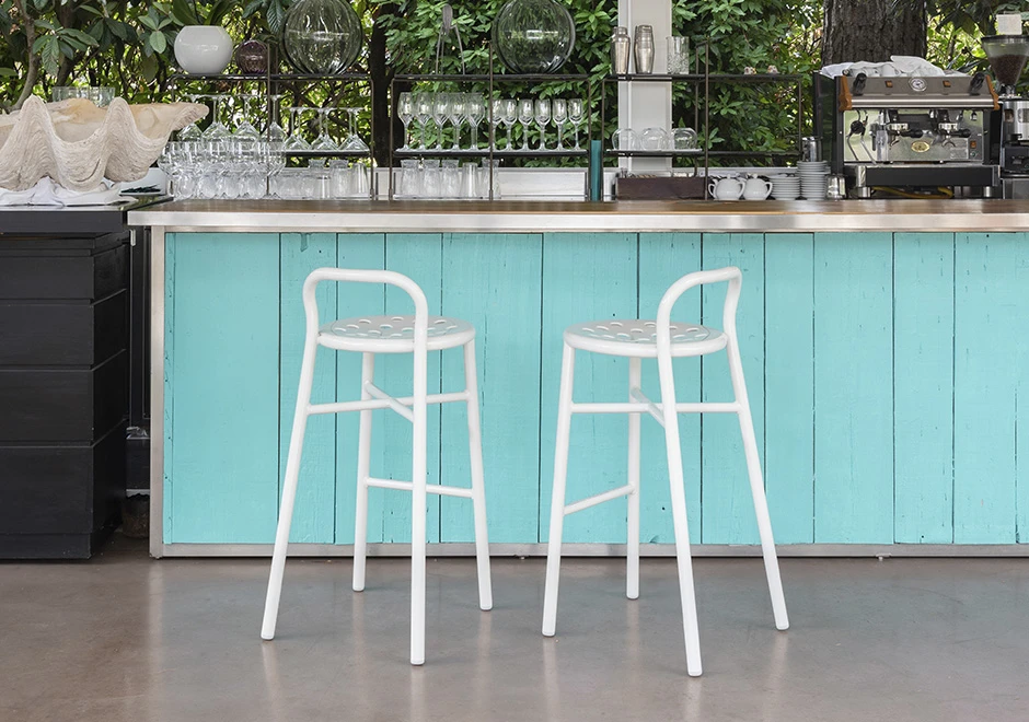 Banco de bar com estrutura de alumínio pintado; assento perfurado em alumínio pintado branco, visto de costas em uma area externa.