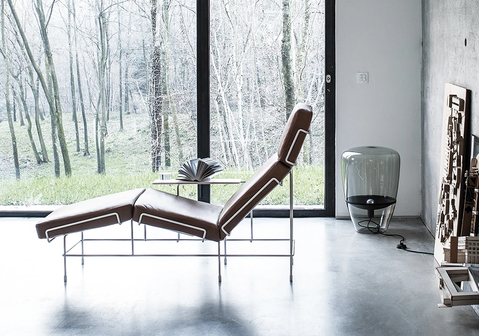 Chaise-longue com estrutura tubular de aço com pintura eletrostática, almofada de assento de poliuretano expandido e capa removível de couro marrom, visto de lado em uma sala moderna.