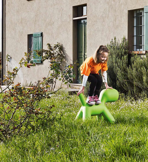 Banco de design em formato de cachorro em polietileno verde, visto de lado, em uma area externa, com uma criança sobre ele.