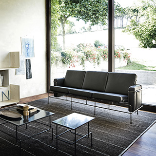 Sofá modular, com estrutura em aço e almofadas em espuma, em tecido cinza, visto de frente em uma sala de parede de vidro.