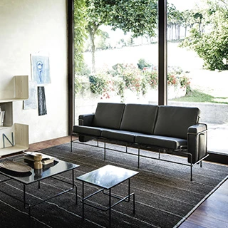 Sofá modular, com estrutura em aço e almofadas em espuma, em tecido cinza, visto de frente em uma sala de parede de vidro.