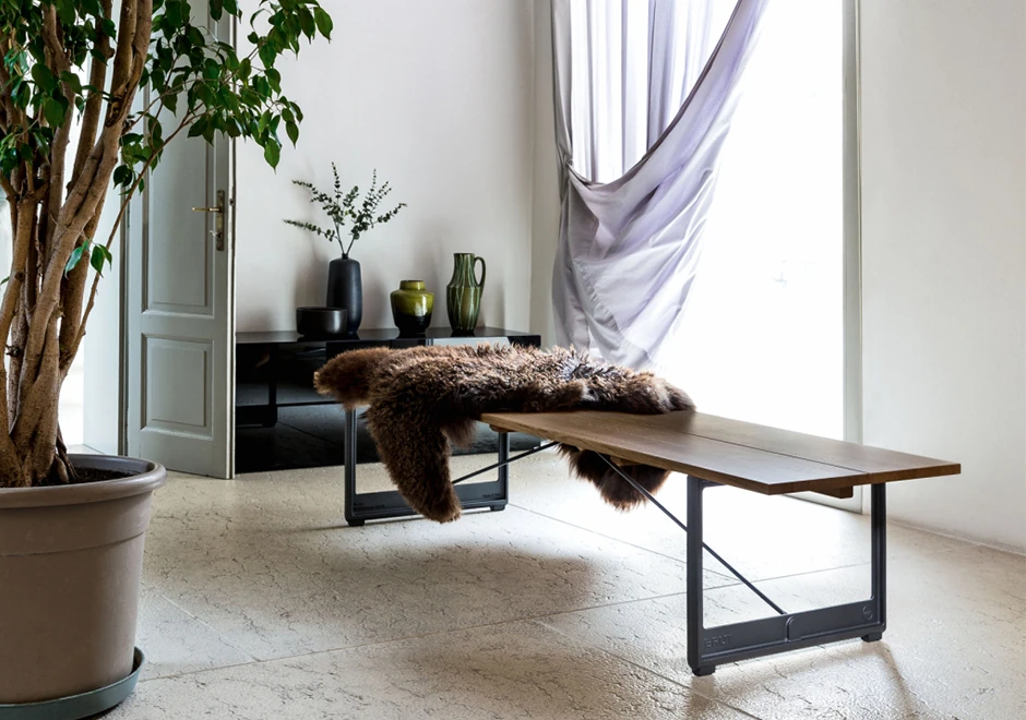 Banco de madeira com estrutura de ferro fundido e assento em Carvalho, com uma pelagem em cima, visto de lado em uma sala moderna.