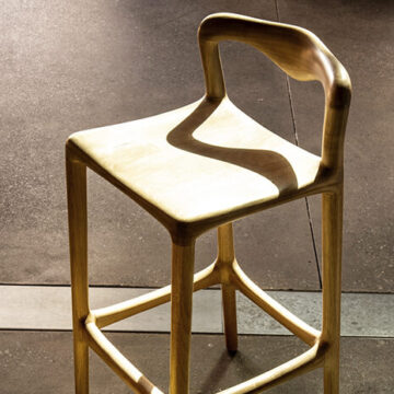 Banco de bar de madeira Tauari natural, visto de lado em um fundo neutro.