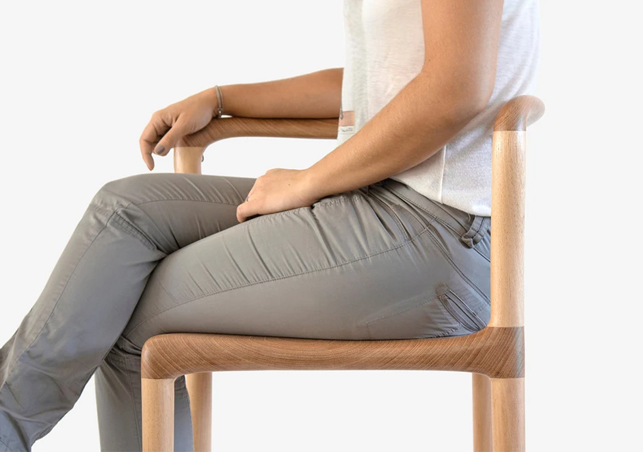 Pessoa sentada em cadeira tripé de madeira Tauari natural, visto de lado, em um fundo branco.