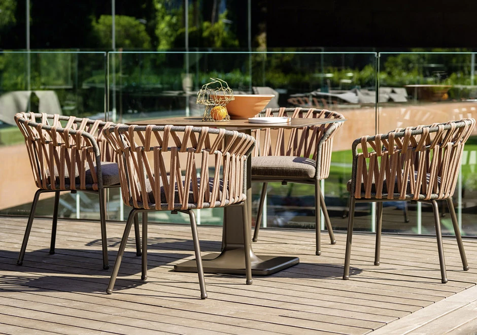 Quatro cadeiras assinadas com estrutura de alumínio com pintura eletrostática; encosto com fita em tecido impermeável trançada à mão. Almofada de assento estofada em tecido marrom, visto de frente em uma area externa.