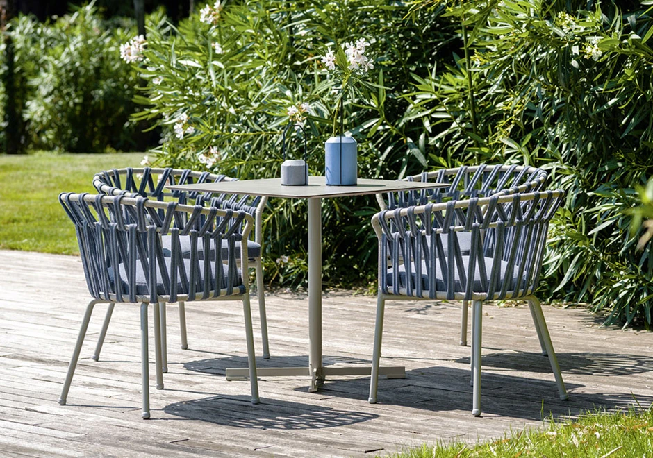 Quatro cadeiras assinadas com estrutura de alumínio com pintura eletrostática; encosto com fita em tecido impermeável trançada à mão. Almofada de assento estofada em tecido azul, visto de frente em uma area externa.