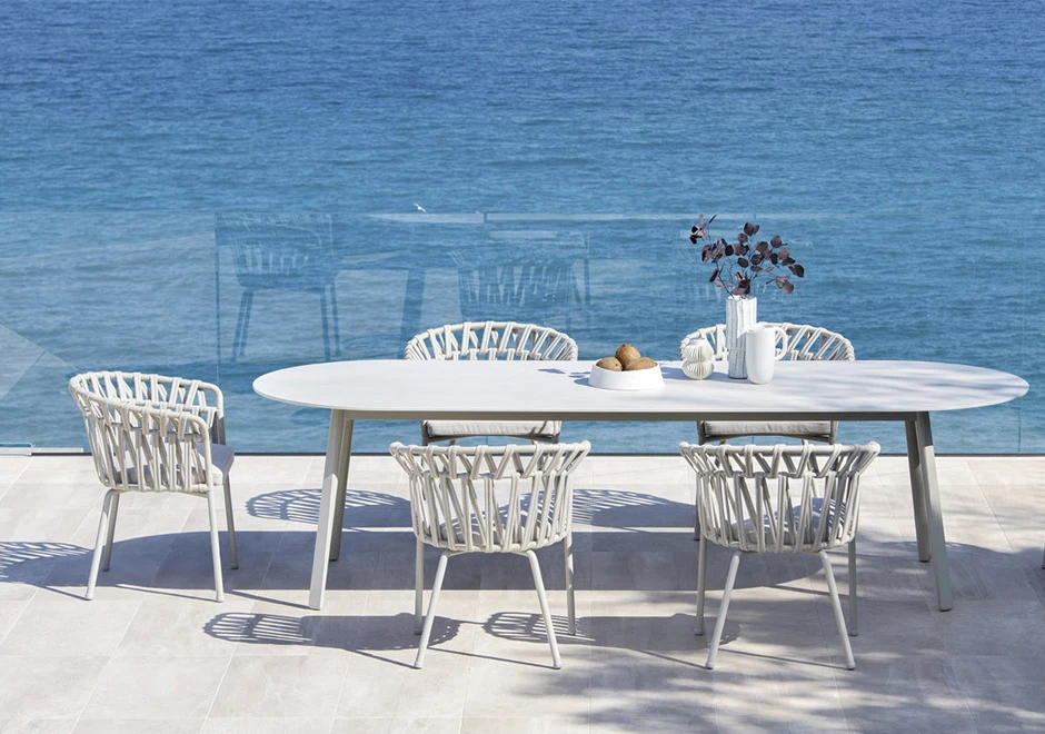 Quatro cadeiras assinadas com estrutura de alumínio com pintura eletrostática; encosto com fita em tecido impermeável trançada à mão. Almofada de assento estofada em tecido branco, visto de frente em uma area externa.