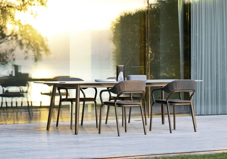 Cinco cadeiras de design com estrutura de alumínio e encosto de fibra sintética trançada a mão na cor bege, em volta de uma mesa em uma area externa.