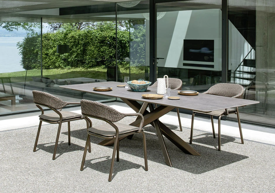 Quatro cadeiras de design com estrutura de alumínio e encosto de fibra sintética trançada a mão na cor bege, em volta de uma mesa, visto de lado, em uma area externa.