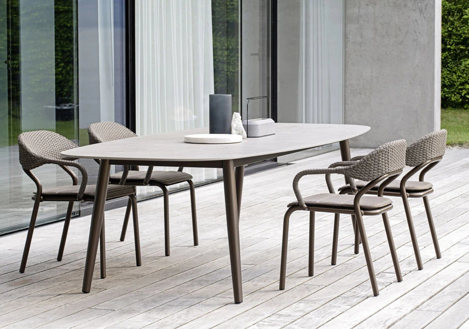 Quatro cadeiras de design com estrutura de alumínio e encosto de fibra sintética trançada a mão na cor bege, em volta de uma mesa, visto de frente, em uma area externa.