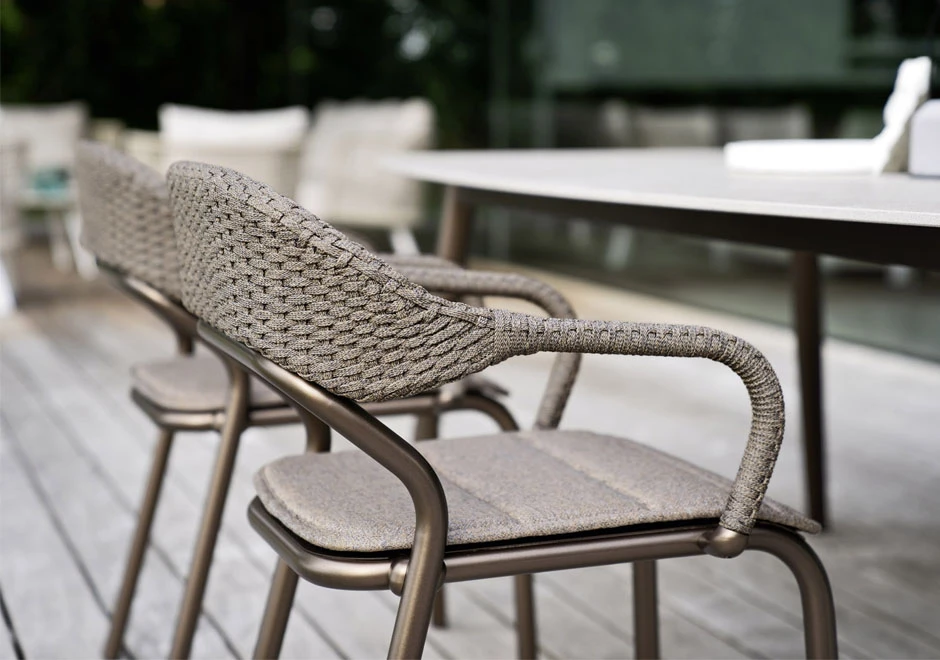Detalhe de cadeiras de design com estrutura de alumínio e encosto de fibra sintética trançada a mão na cor bege, em volta de uma mesa em uma area externa.