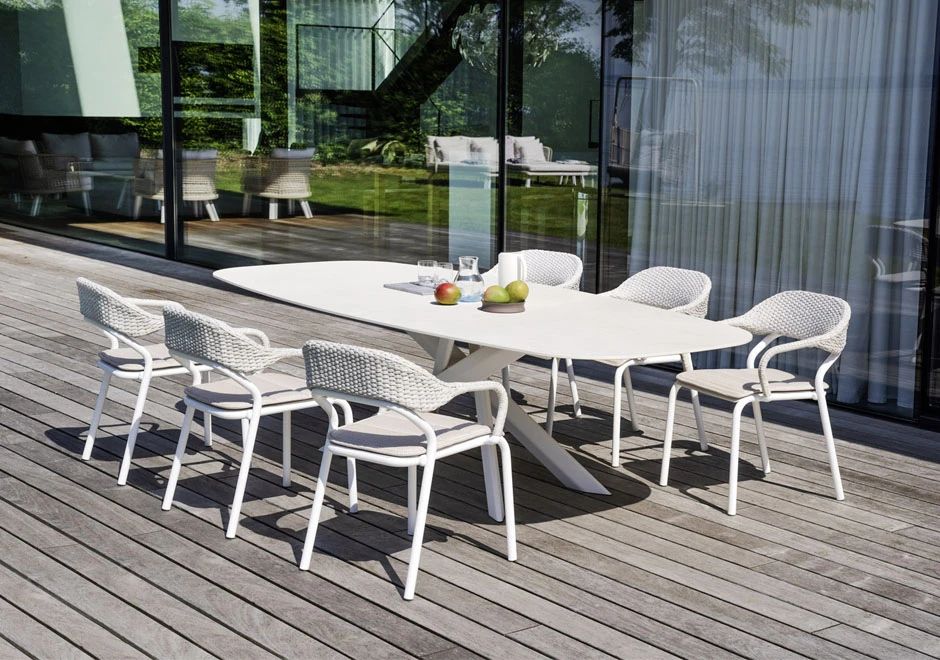 Seis cadeiras de design com estrutura de alumínio e encosto de fibra sintética trançada a mão na cor branco, em volta de uma mesa em uma area externa.