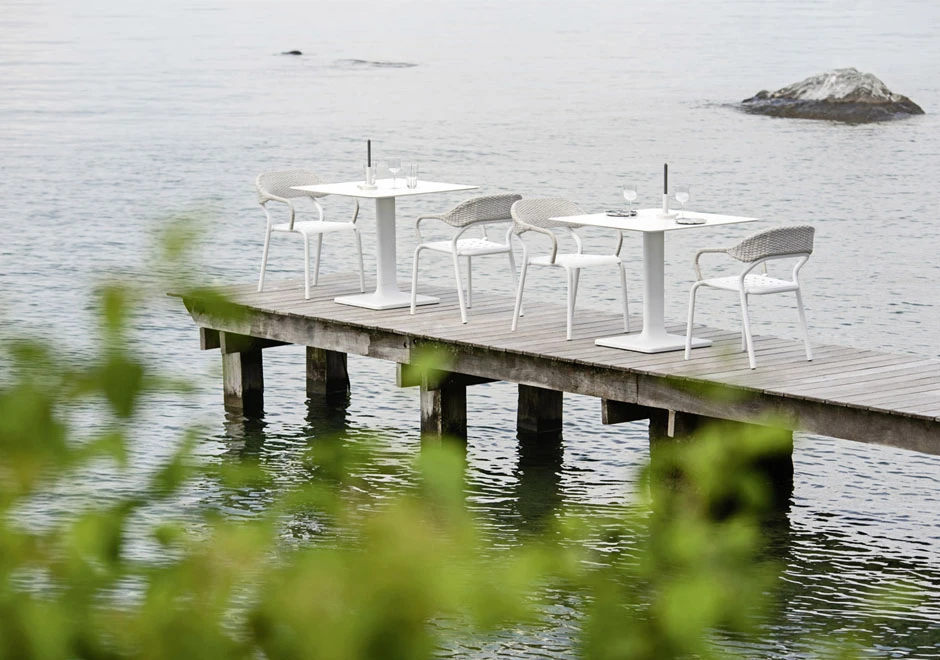 Quatro cadeiras de design com estrutura de alumínio e encosto de fibra sintética trançada a mão na cor branco, em volta de duas mesas em uma area externa.