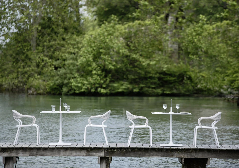 Quatro cadeiras de design com estrutura de alumínio e encosto de fibra sintética trançada a mão na cor branco, em volta de duas mesas, visto de lado, em uma area externa.