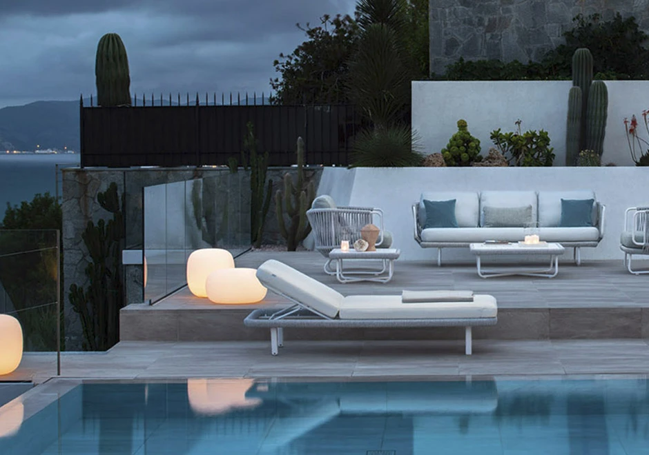 Chaise longue com estrutura e rodinhas em alumínio pintado, corda de fibra sintética de cânhamo, almofada em tecido branco, visto de de lado em uma area externa de piscina.