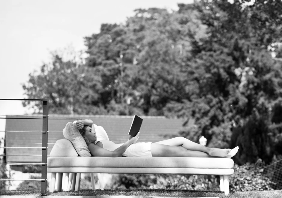 Chaise longue com estrutura de alumínio com pintura eletrostática, pés em alumínio pintado ou madeira Iroko natural, tecido na cor bege, visto de lado, com uma pessoa deitada, em uma area externa.