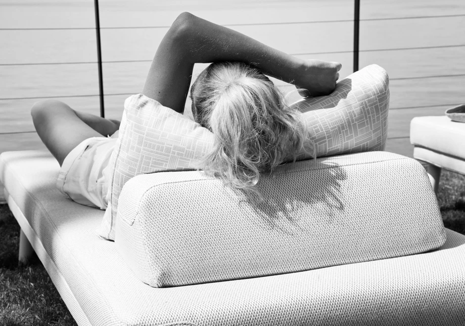 Chaise longue com estrutura de alumínio com pintura eletrostática, pés em alumínio pintado ou madeira Iroko natural, tecido na cor bege, visto de costas, com uma pessoa deitada, em uma area externa.