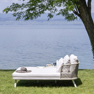 Chaise Daybed com estrutura de alumínio com pintura eletrostática branca; encosto em fibra sintética trançada à mão, pés de alumínio pintado branco, visto de lado em uma area externa.