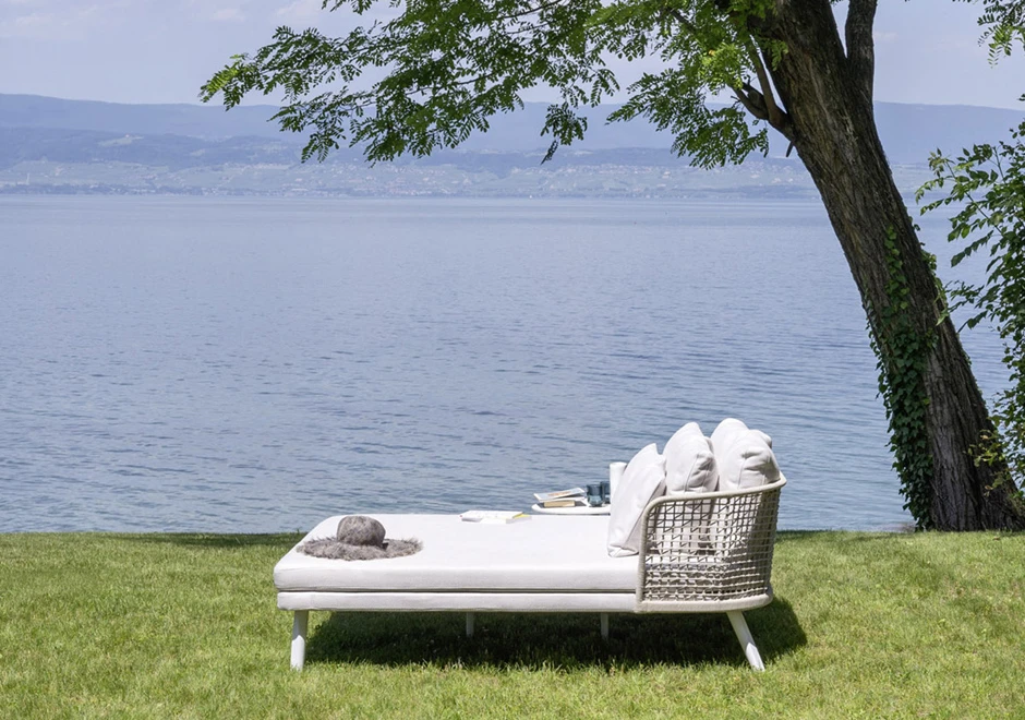 Chaise Daybed com estrutura de alumínio com pintura eletrostática branca; encosto em fibra sintética trançada à mão, pés de alumínio pintado branco, visto de lado em uma area externa.