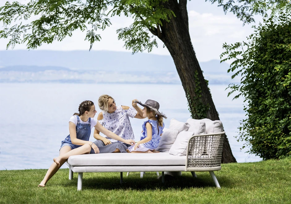 Chaise Daybed com estrutura de alumínio com pintura eletrostática branca; encosto em fibra sintética trançada à mão, pés de alumínio pintado branco, visto de lado com algumas pessoas sentadas, em uma area externa.