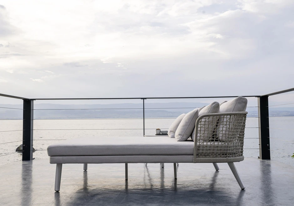 Chaise Daybed com estrutura de alumínio com pintura eletrostática branca; encosto em fibra sintética trançada à mão, pés de alumínio pintado branco, visto de lado em uma area externa de lago.
