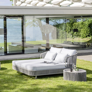 Chaise-longue com estrutura de alumínio com pintura eletrostática, revestida com fibras sintéticas trançadas à mão, dois pés com rodinhas e estofado com espuma branca, visto de lado em uma area externa.