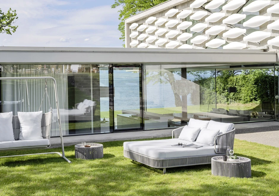 Chaise-longue com estrutura de alumínio com pintura eletrostática, revestida com fibras sintéticas trançadas à mão, dois pés com rodinhas e estofado com espuma branca, visto de lado em uma area externa.