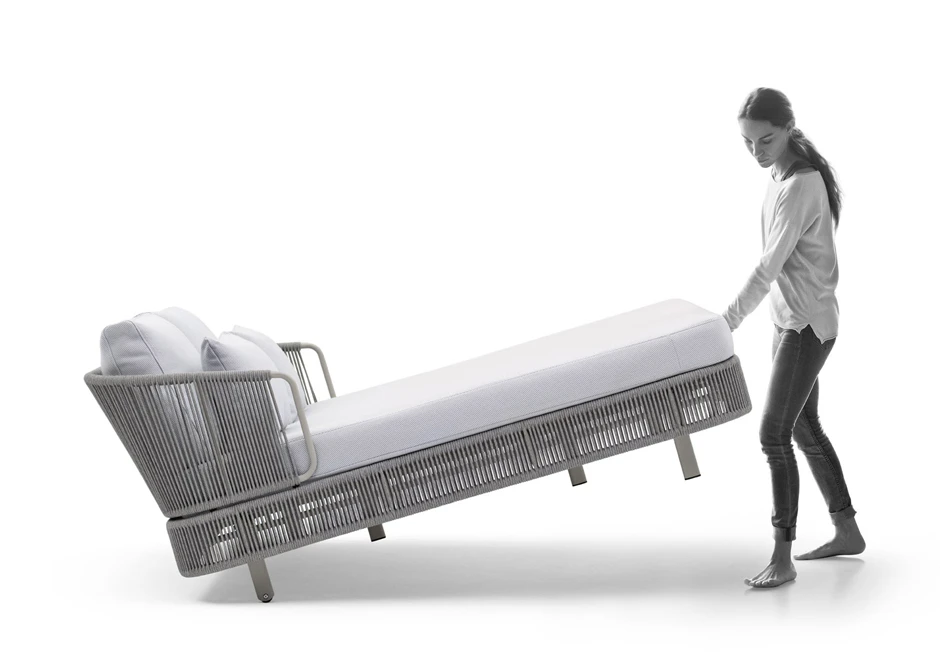 Chaise-longue com estrutura de alumínio com pintura eletrostática, revestida com fibras sintéticas trançadas à mão, dois pés com rodinhas e estofado com espuma branca, visto de lado, com uma pessoa carregando ele, em um fundo branco.