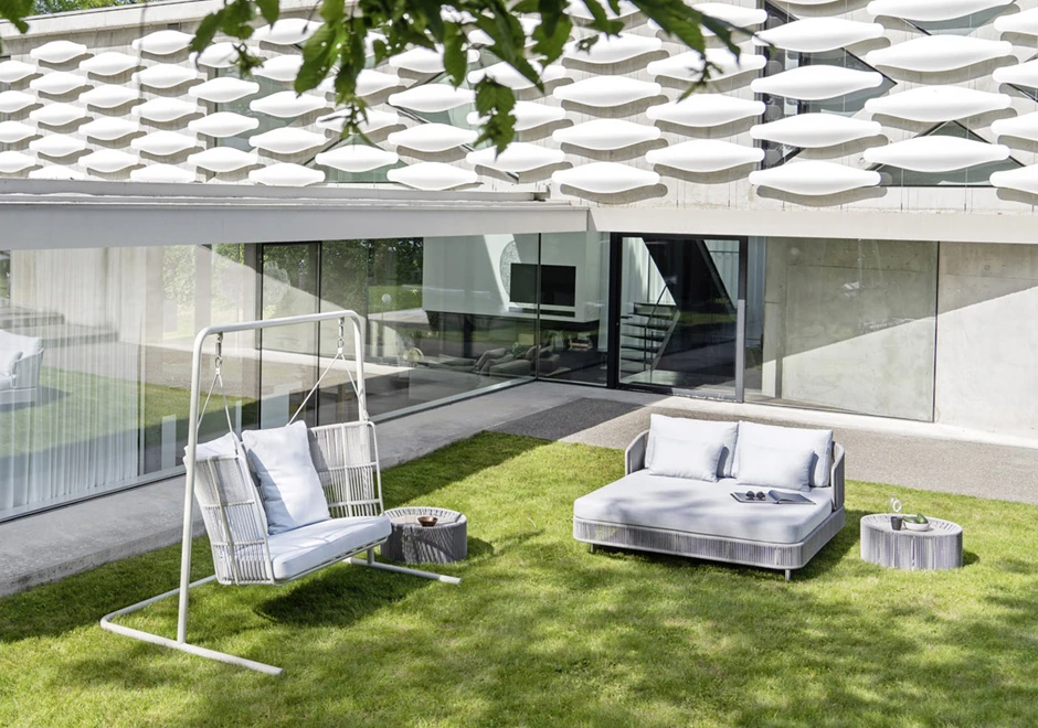 Chaise-longue com estrutura de alumínio com pintura eletrostática, revestida com fibras sintéticas trançadas à mão, dois pés com rodinhas e estofado com espuma branca, visto de frente em uma area externa.