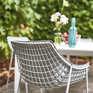 Cadeira de design com estrutura de aço cromado ou pintado e tecido branco, vista de costas, em uma area externa.