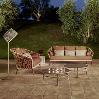 Sofá com encosto em fita trançada à mão, estrutura de alumínio, estofado na cor rosa e almofadas brancas, visto de frente em uma area externa, a noite.