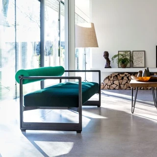 Sofá contemporâneo com estrutura em ferro fundido, e estofado em espuma, verde claro no encosto e verde escuro no assento, visto de lado em uma sala moderna de janelas grandes e sol batendo no interior.