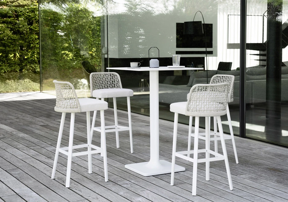 Quatro bancos de design, com estrutura em alumínio com pintura branca. Assento revestido em tecido e encosto com estrutura de alumínio com acabamento em corda trançada à mão, branco, ao redor de uma mesa em uma area externa.