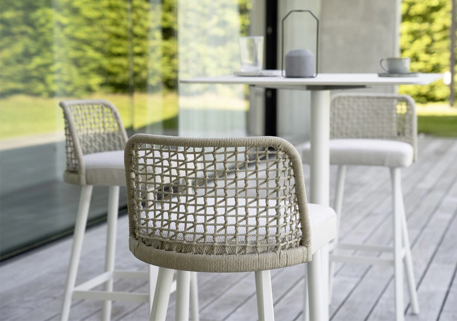 Encosto de bancos de design, com estrutura em alumínio com pintura branca. Assento revestido em tecido e encosto com estrutura de alumínio com acabamento em corda trançada à mão, branco, ao redor de uma mesa em uma area externa.