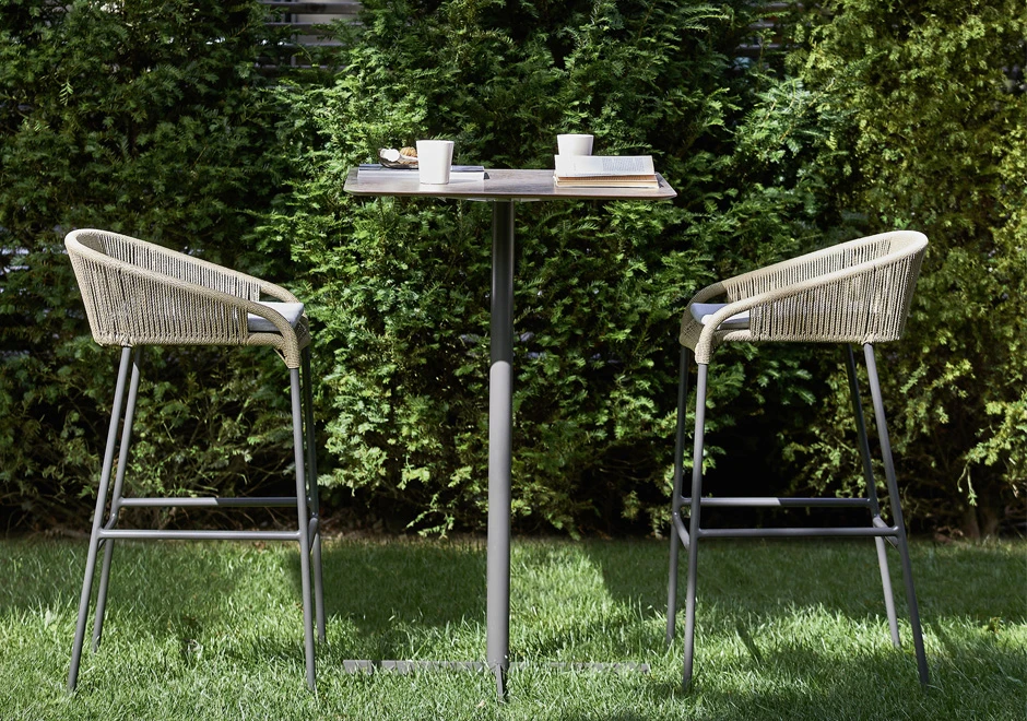 Dois bancos de alumínio com assento e encosto revestidos com fibra sintética trançada a mão, cinza, em frente a uma mesa, em uma area externa.