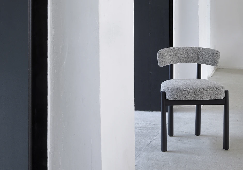Uma cadeira assinada com estrutura freixo maciço tingido de preto e assento e encosto estofados em tecido cinza, vista de frente em uma sala vazia.