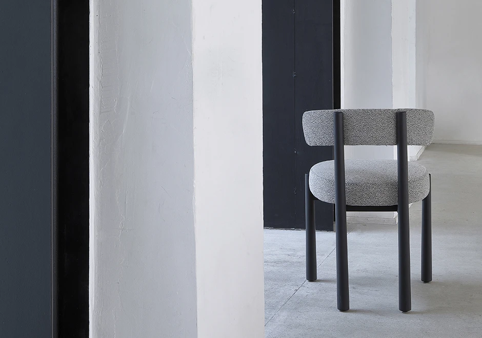 Uma cadeira assinada com estrutura freixo maciço tingido de preto e assento e encosto estofados em tecido cinza, vista de costas em uma sala vazia.