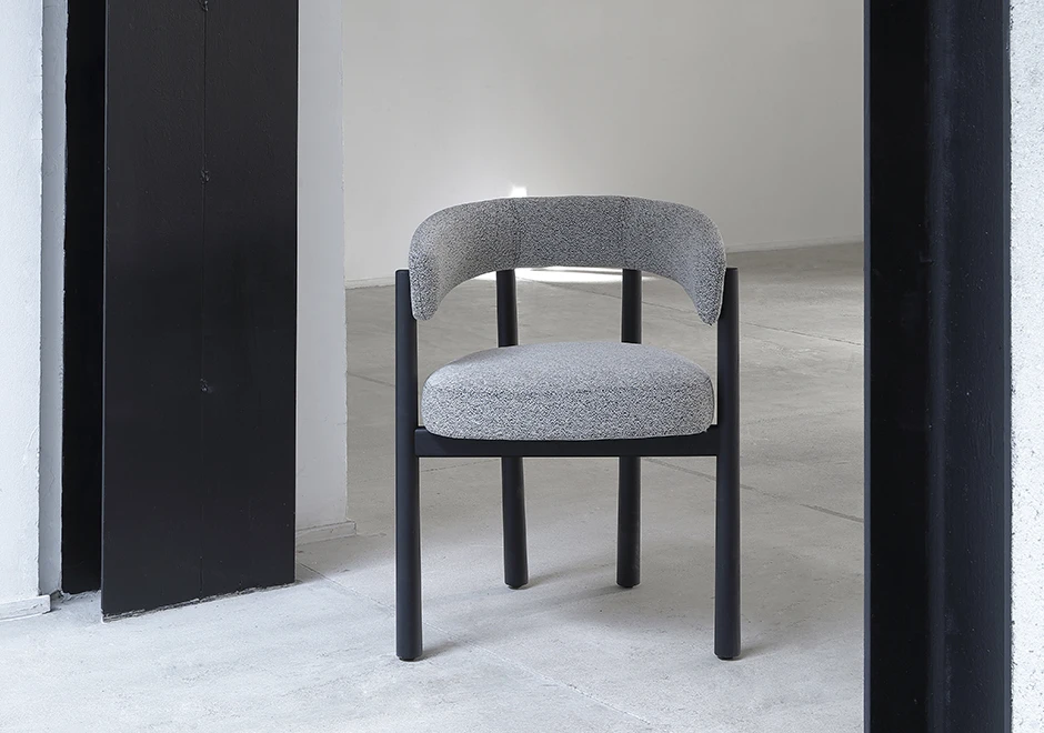 Uma cadeira assinada com estrutura freixo maciço tingido de preto e assento e encosto estofados em tecido cinza, vista de frente em uma sala vazia.