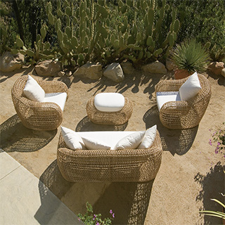 Imagem vista de cima, de um sofá de dois lugares de tecido branco, almofadas brancas e estrutura em fibra sintética trançada, duas poltronas de de tecido branco, almofadas brancas e estrutura em fibra sintética trançada e um pufe ao centro com as mesmas caracteristicas. Imagem em uma área externa de jardim.