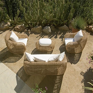 Imagem vista de cima, de um sofá de dois lugares de tecido branco, almofadas brancas e estrutura em fibra sintética trançada, duas poltronas de de tecido branco, almofadas brancas e estrutura em fibra sintética trançada e um pufe ao centro com as mesmas caracteristicas. Imagem em uma área externa de jardim.