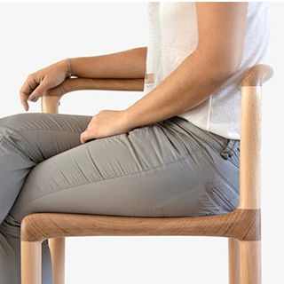 Pessoa sentada em cadeira tripé de madeira Tauari natural, visto de lado, em um fundo branco.